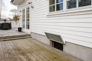 Bakside av enebolig i Kristiansand med terrasse og inngang til krypkjeller, motiv viser område ved stue hvor lukt er rapportert.